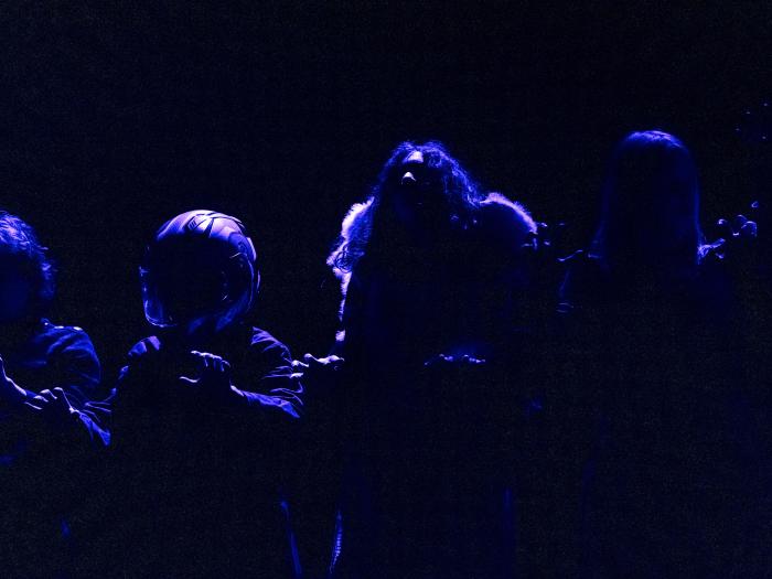 Mørkt blåt baglys på scene med fire skikkelser. Blandt andet en med vinger og en med en styrthjelm på