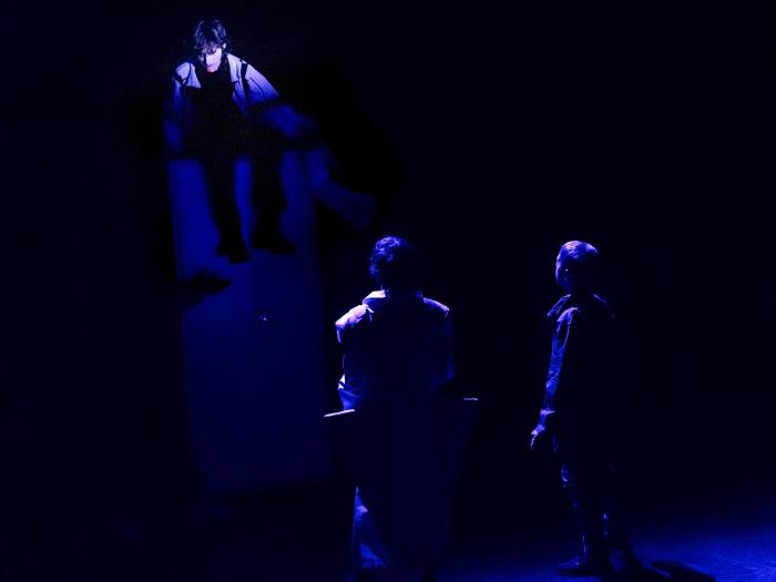 Mørk scene og lavt blåt lys. To børn kigger op på et tredje barn, som sidder højt oppe på et skab