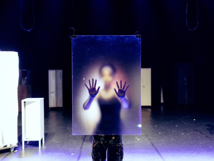 Mørk, blå scene med skabe rundt omkring. Et barn bag en plexiglas-plade med begge hænder på den