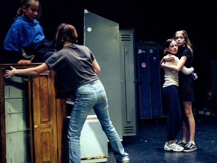 To børn krammer hinanden på en mørk scene. Et andet barn sidder på et lavt skab som skubbes af et fjerde barn